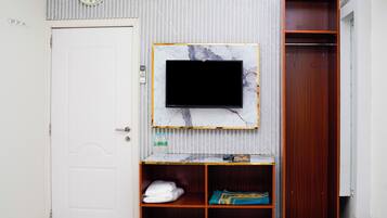 A room with a white door, a clock, a TV on a marble-framed wall, a wooden wardrobe, and a shelf with towels and a book.