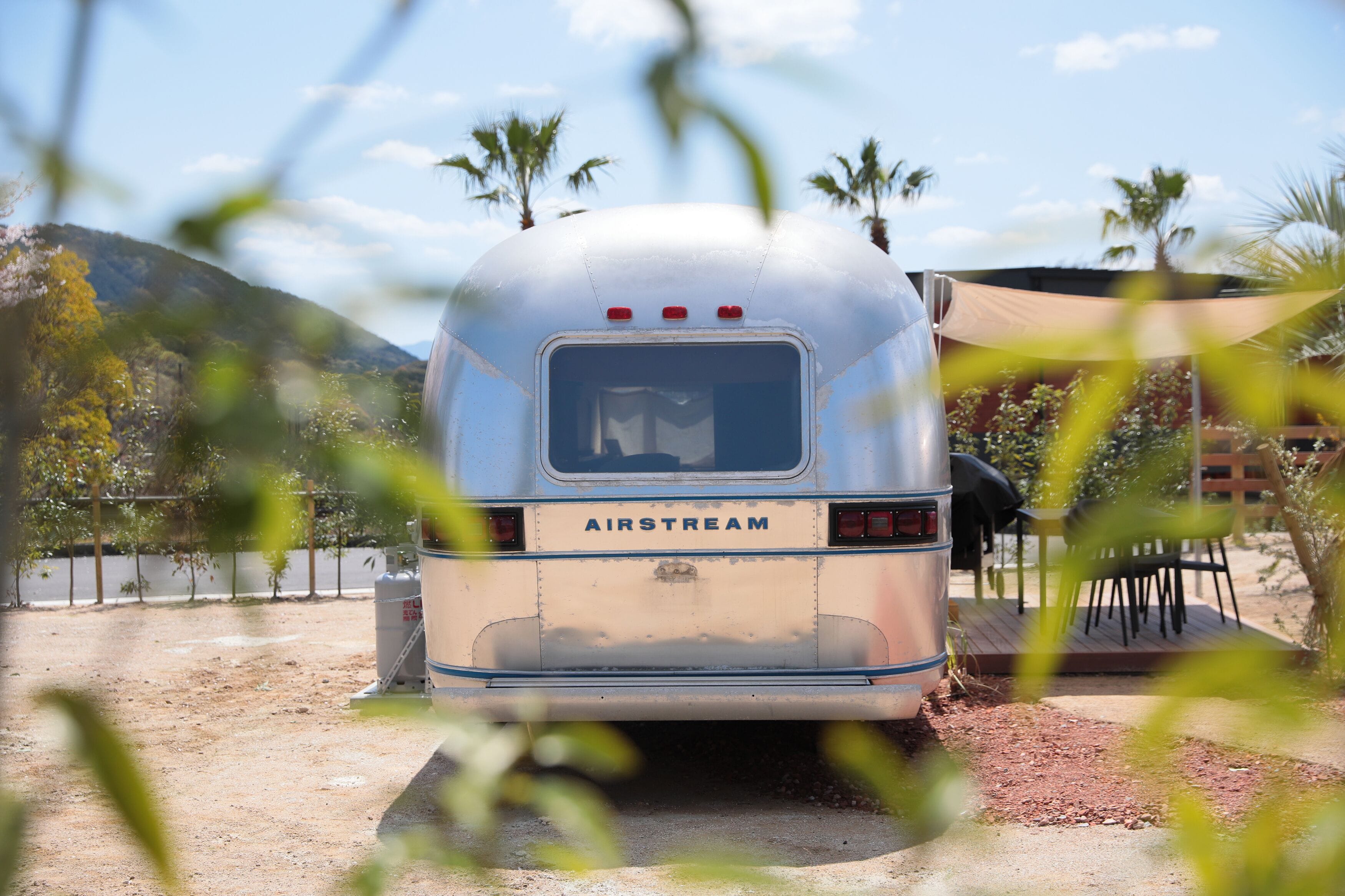 Uma caravana AIRSTREAM antiga estacionada em área externa ensolarada, com palmeiras e um deck de madeira.