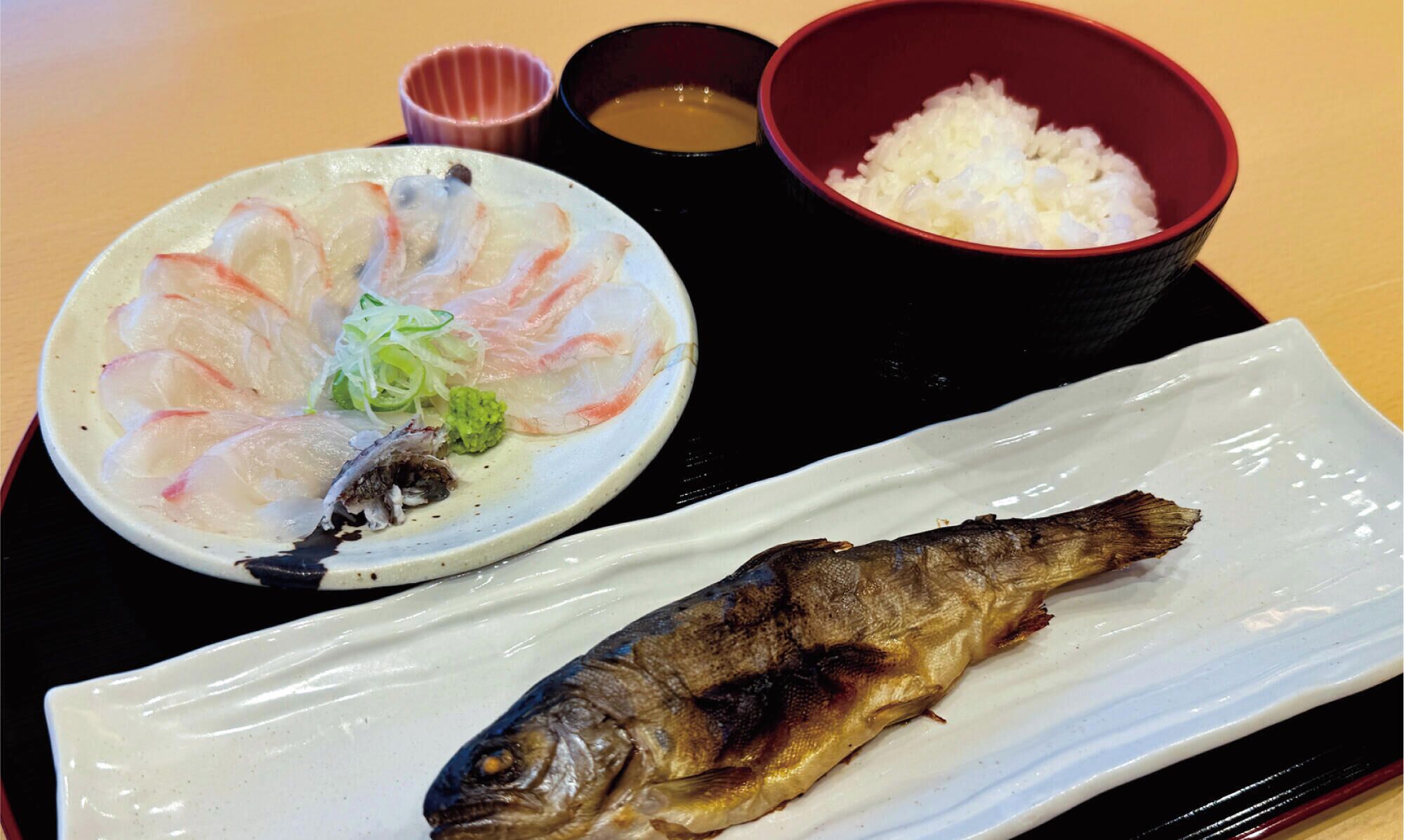 Eine Platte mit Sashimi, eine Schüssel Reis und ein gegrillter Fisch auf einem Tablett.