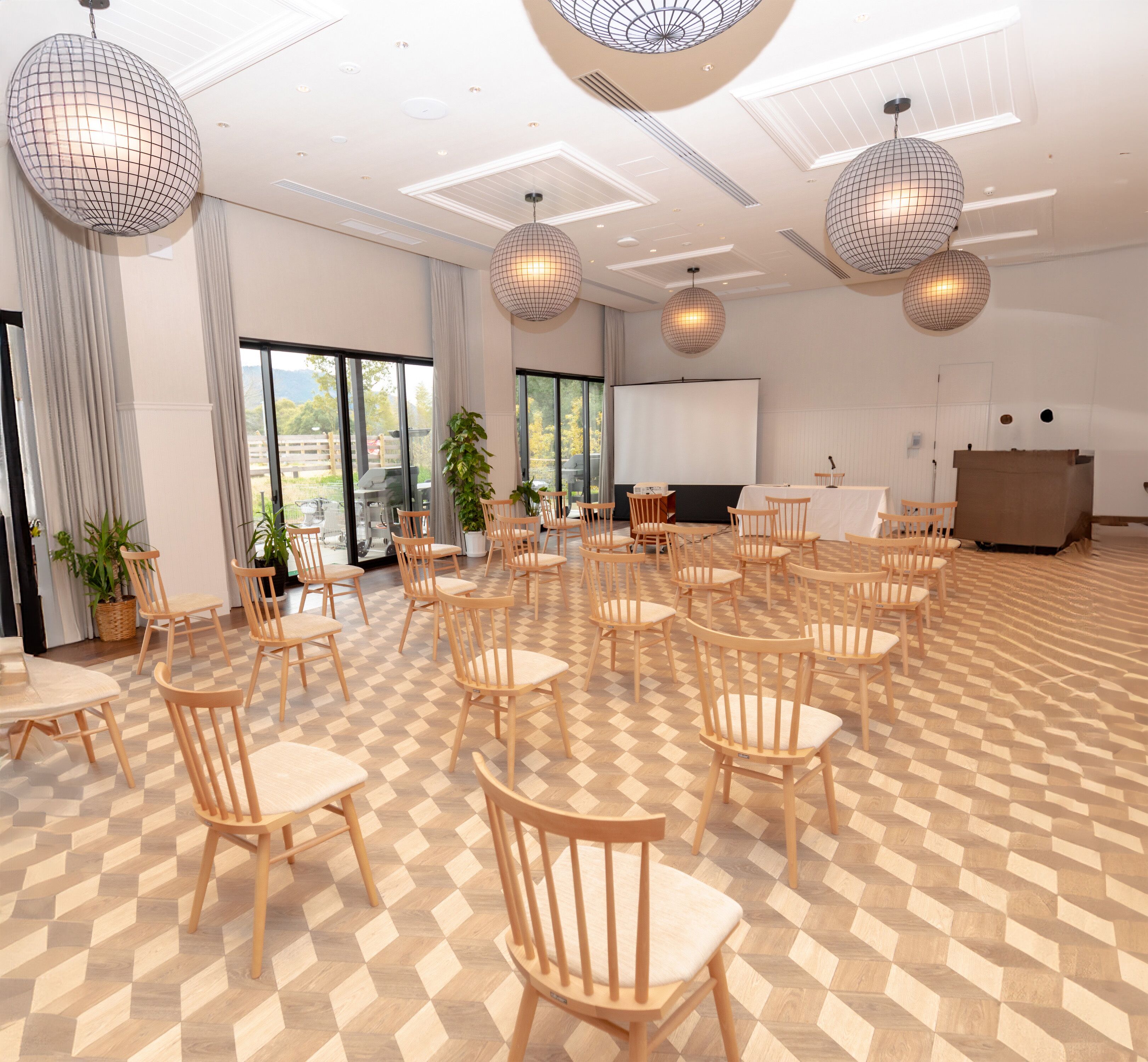 A spacious room with wooden chairs and tables, large windows, and a projector screen.