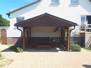 Un’area salotto esterna coperta con una panchina e un tavolo in legno, un piccolo giardino e un edificio bianco con finestre sullo sfondo.
