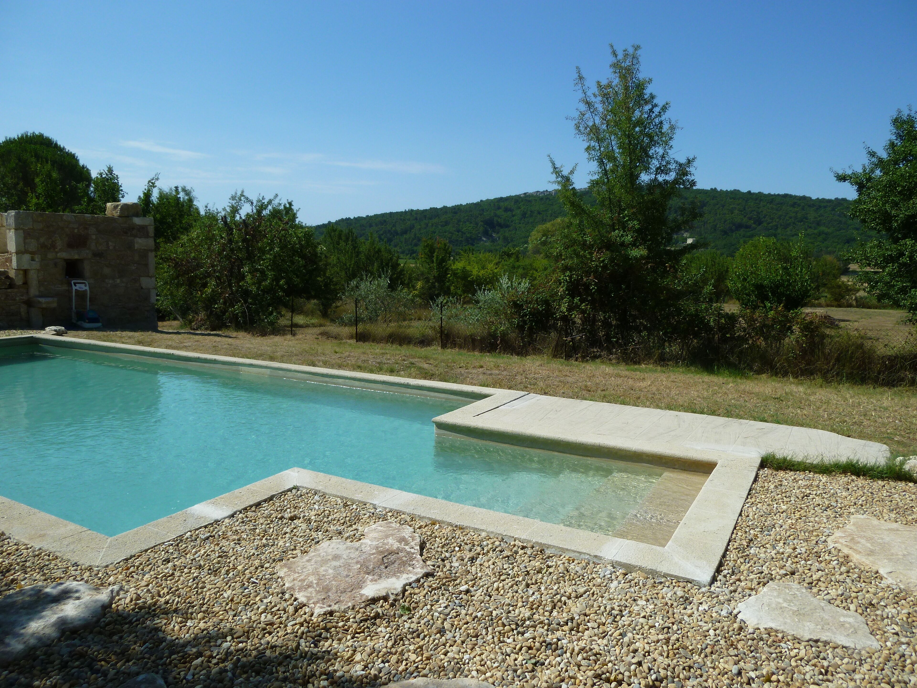Una piscina rectangular con borde de piedras, rodeada por un muro de piedra y vegetación.