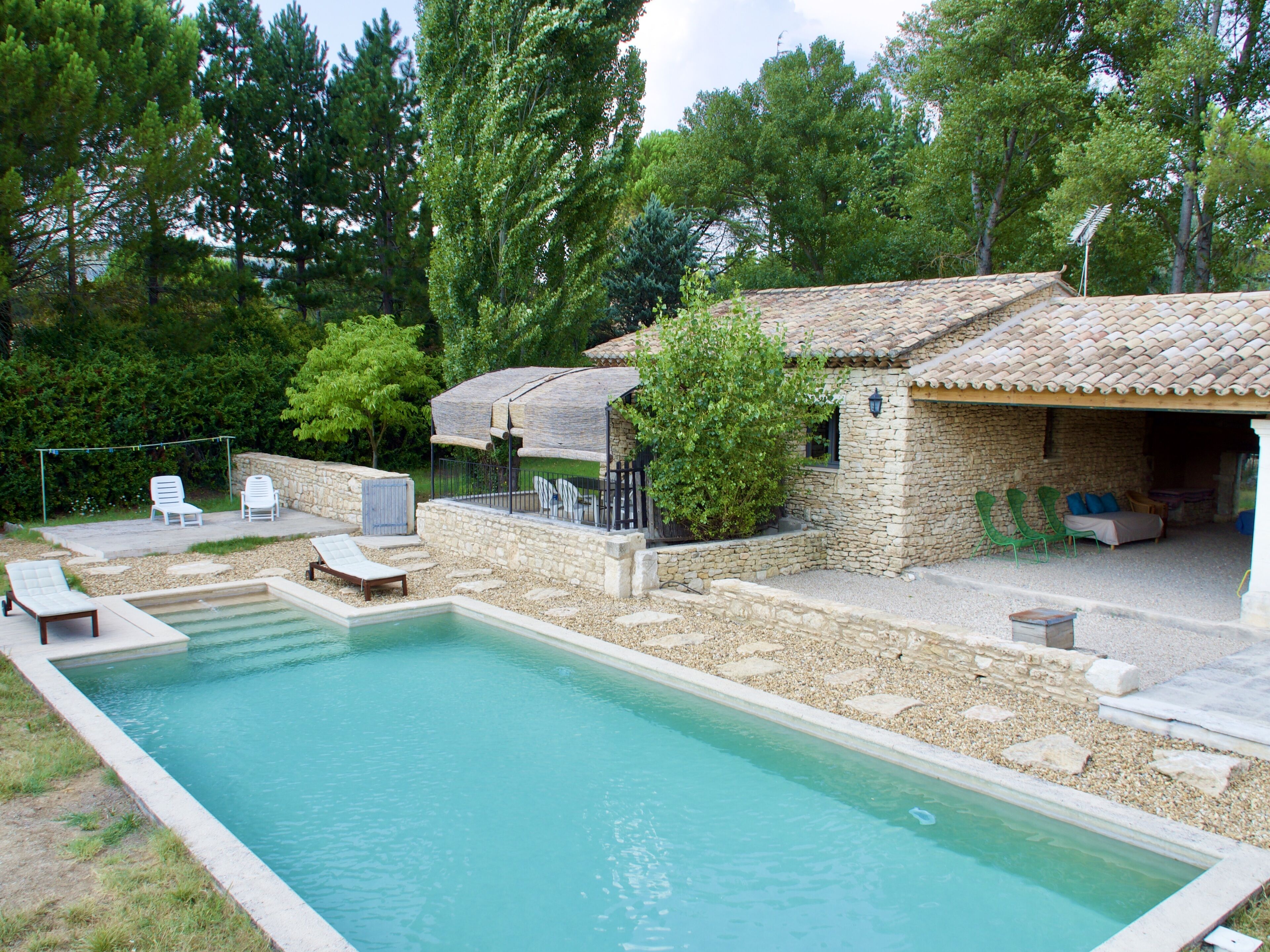 A swimming pool with lounge chairs, a stone building with a tiled roof, and surrounding greenery.