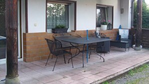An outdoor patio with a table and chairs, a potted plant, and a window with a view of trees.