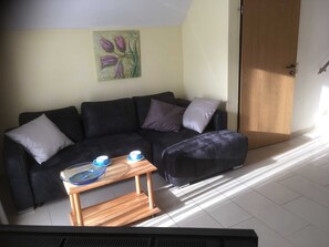 Uma sala de estar com um sofá preto, uma mesa de centro de madeira com duas xícaras e um quadro de flores roxas na parede.