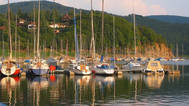 Ein Yachthafen mit verschiedenen Schiffen, umgeben von einer hügeligen Landschaft und Häusern im Hintergrund.
