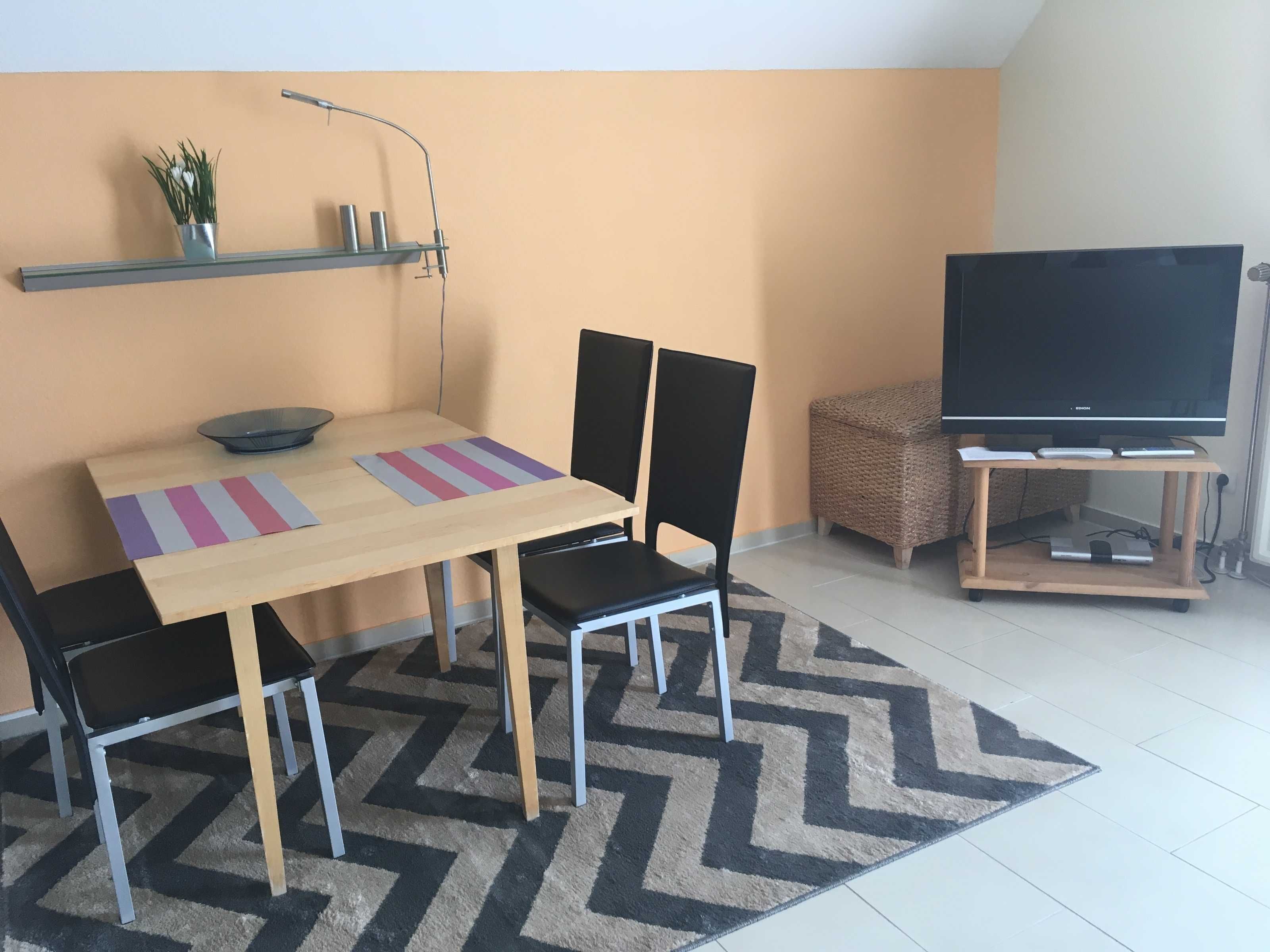 Un espacio habitable compacto con una mesa de comedor de madera, cuatro sillas, un televisor sobre un mueble bajo, un estante con adornos y una alfombra estampada.