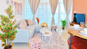 Un salon moderne comprenant un canapé, un pouf, une table basse et un coin repas avec une table et des chaises. On y trouve également un téléviseur posé sur un meuble et une fenêtre avec des rideaux.