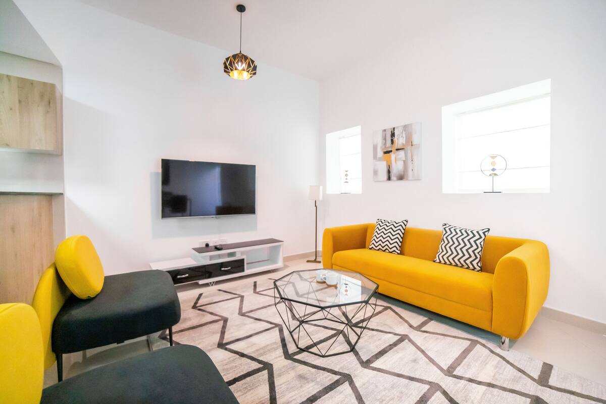 Een moderne woonkamer met een gele bank, zwarte en gele fauteuils, een glazen salontafel, een flatscreen televisie en kunstwerken aan de muur.
