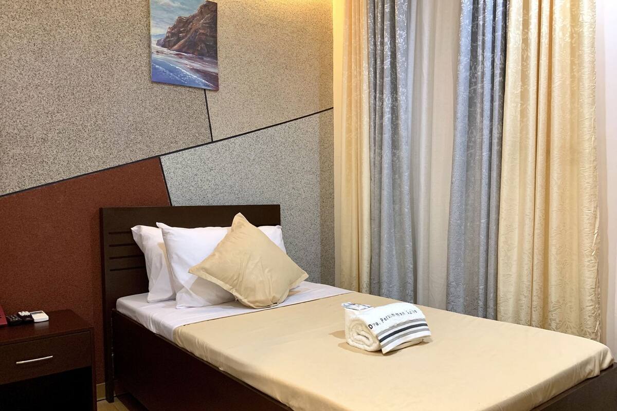A hotel room with a bed, a nightstand, a remote control, and a towel with text.