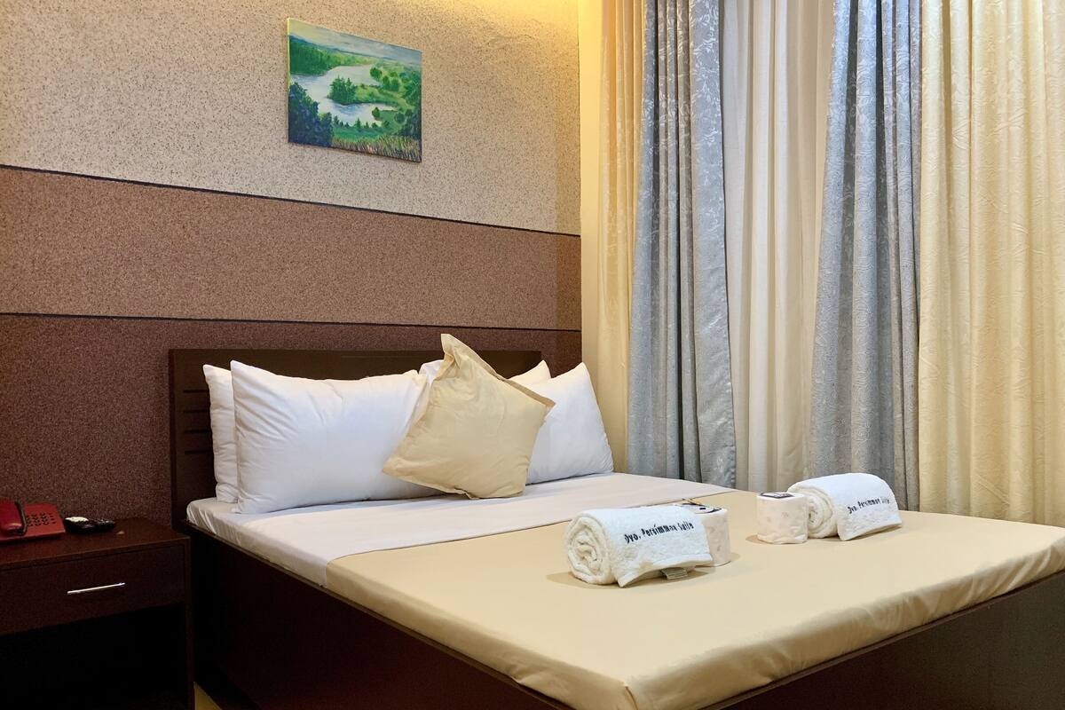 A hotel room with a bed, pillows, a painting on the wall, and curtains.