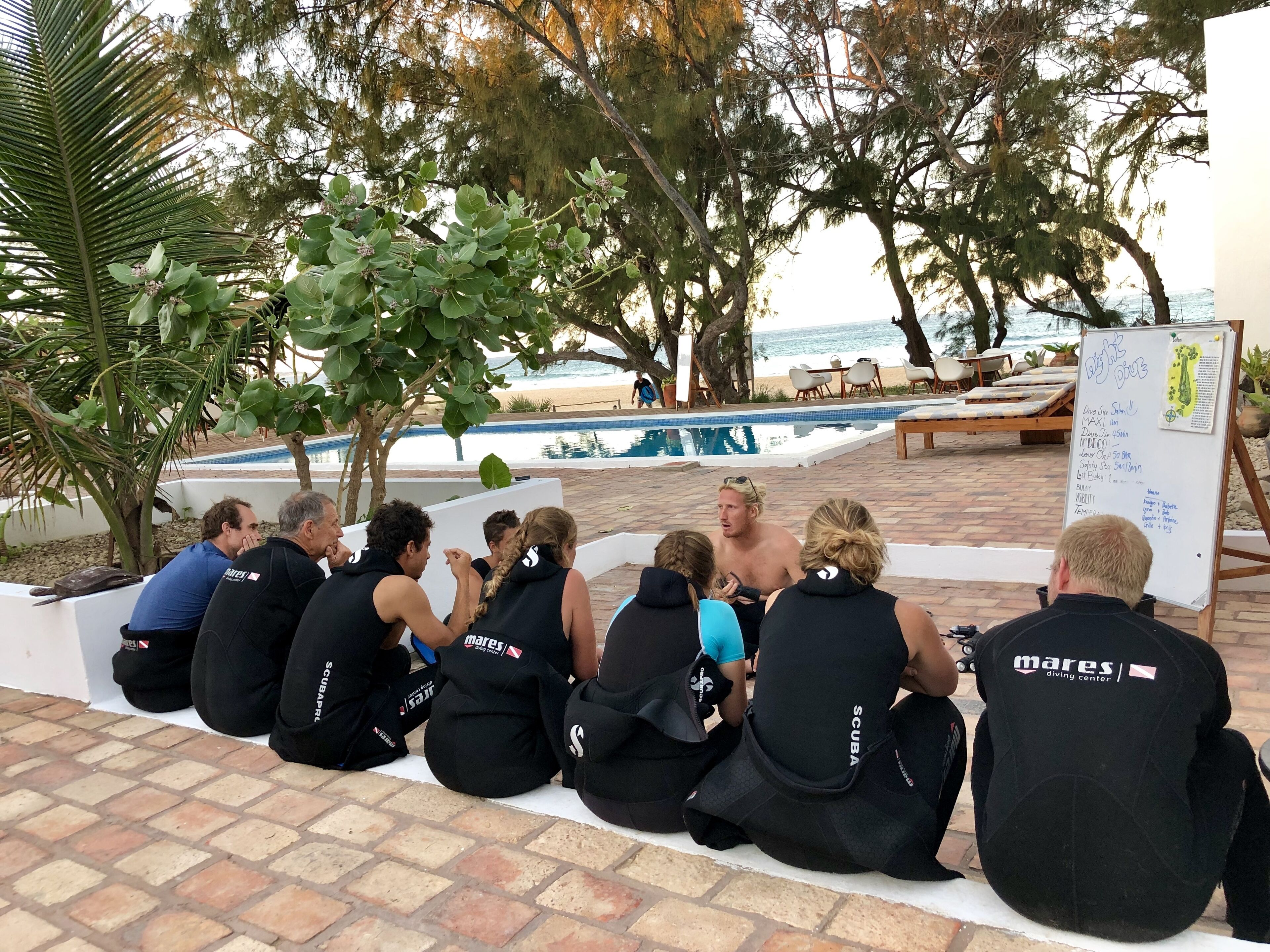 A group of people sitting on a tiled floor near a poolside area with a whiteboard displaying information.