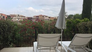 A patio with two white lounge chairs and a grey umbrella, surrounded by greenery and a view of buildings in the background.