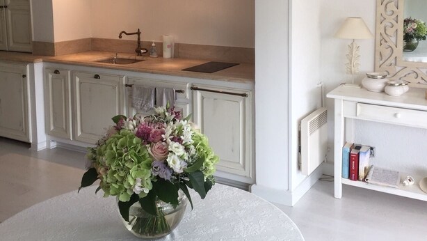A modern kitchen with white cabinetry, a round dining table with a floral centerpiece, and a white console table with a lamp and books.