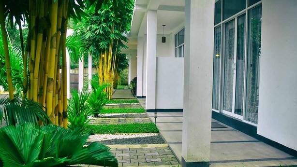 A corridor with a tiled pathway, green plants, and a building with large windows. - Hotel Aram (Unawatuna)