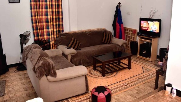 A living room with a brown sofa, a beige armchair, a coffee table, a television on a stand, a fan, and a patterned rug. - Hotel Aram (Unawatuna)