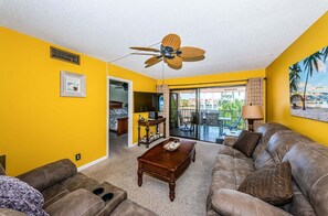 Een woonkamer met gele wanden, een plafondventilator, een bank, een salontafel en uitzicht op een balkon.