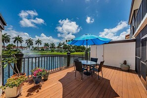 Una terraza de madera con una mesa y sillas, una sombrilla azul y vistas a un cuerpo de agua con palmeras.