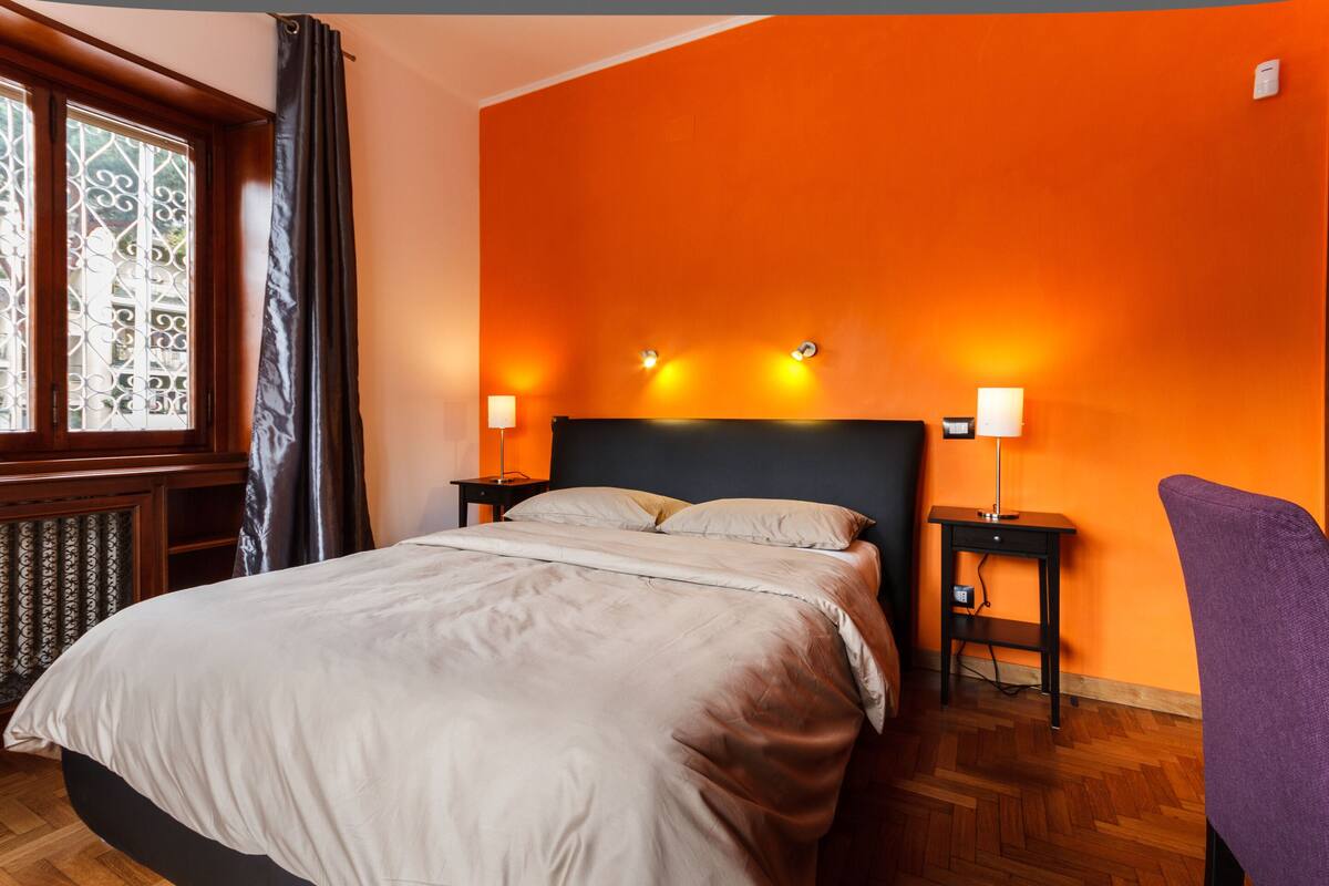 A bedroom with a bed, two bedside tables, a window with curtains, and an orange wall.