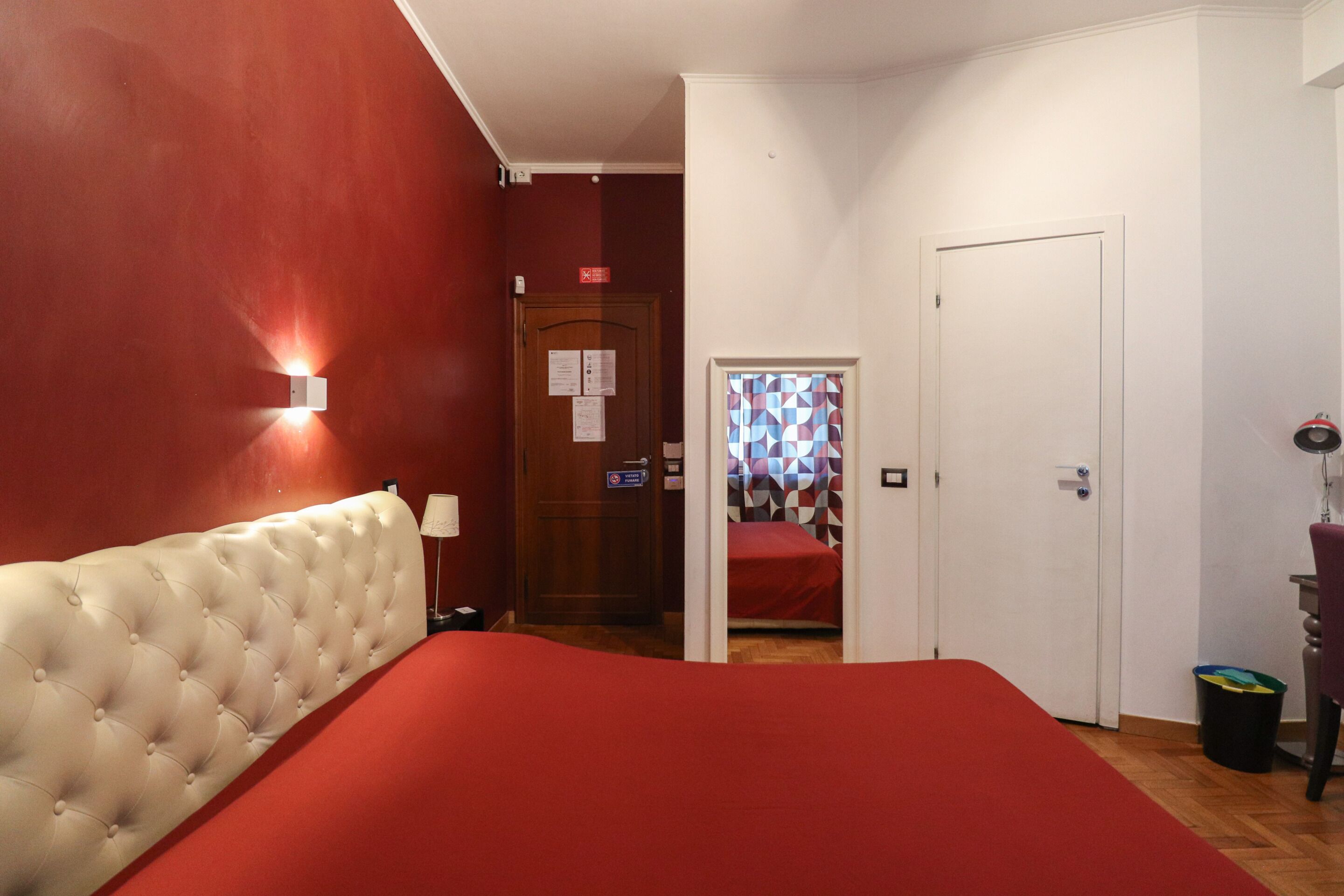 Ein Hotelzimmer mit roter Wand, einem gepolsterten Kopfteil, einer roten Tagesdecke, einer Holztür und einem kleinen Fenster mit einem geometrischen Vorhang.