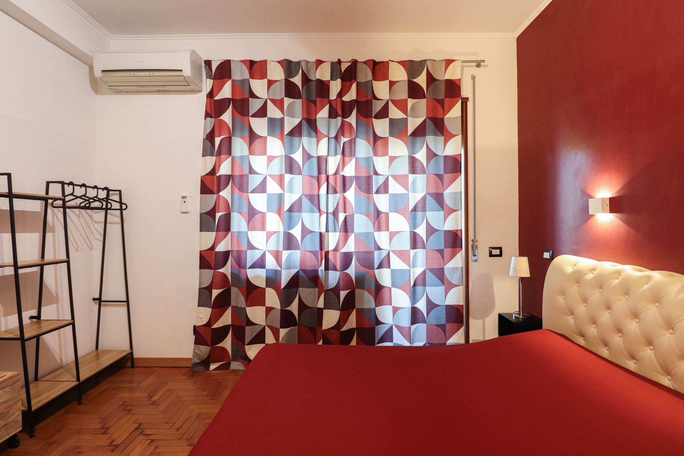 Een slaapkamer met een rode sprei, een gordijn met een geometrisch patroon, een hoofdbord, een nachtkastje met een lamp en een wandlamp.