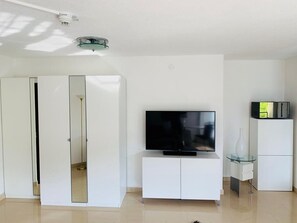 Ett modernt vardagsrum med en platt-TV placerad på ett vitt skåp, ett glasbord och en vit garderob med en spegel.