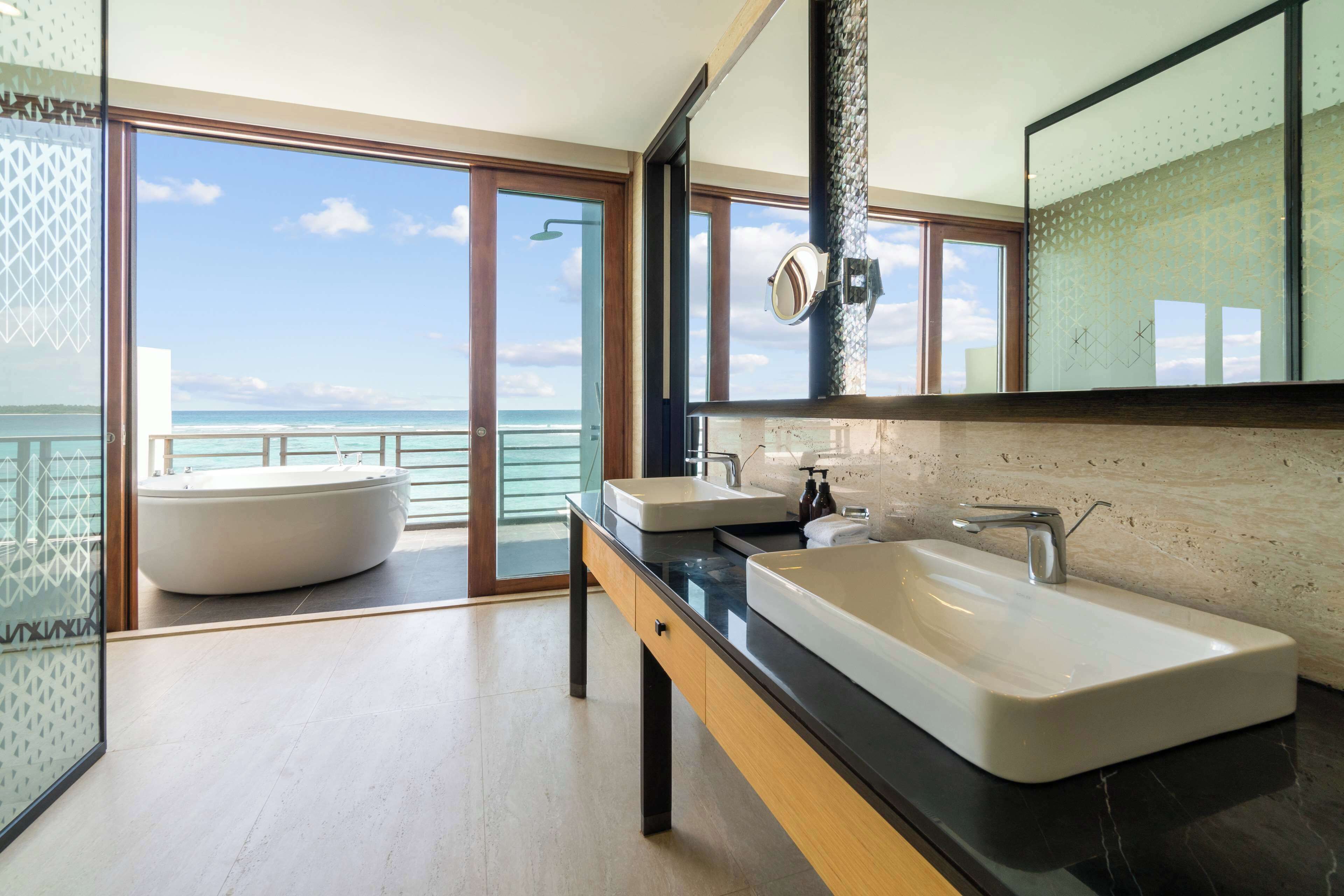 Une salle de bain moderne avec deux vasques, un grand miroir et une vue sur l’océan par une porte vitrée.