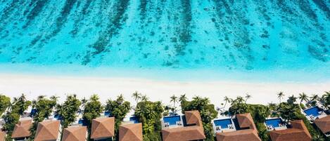 An aerial view of a resort with multiple bungalow-style buildings, each with a pool, surrounded by palm trees and a beach.