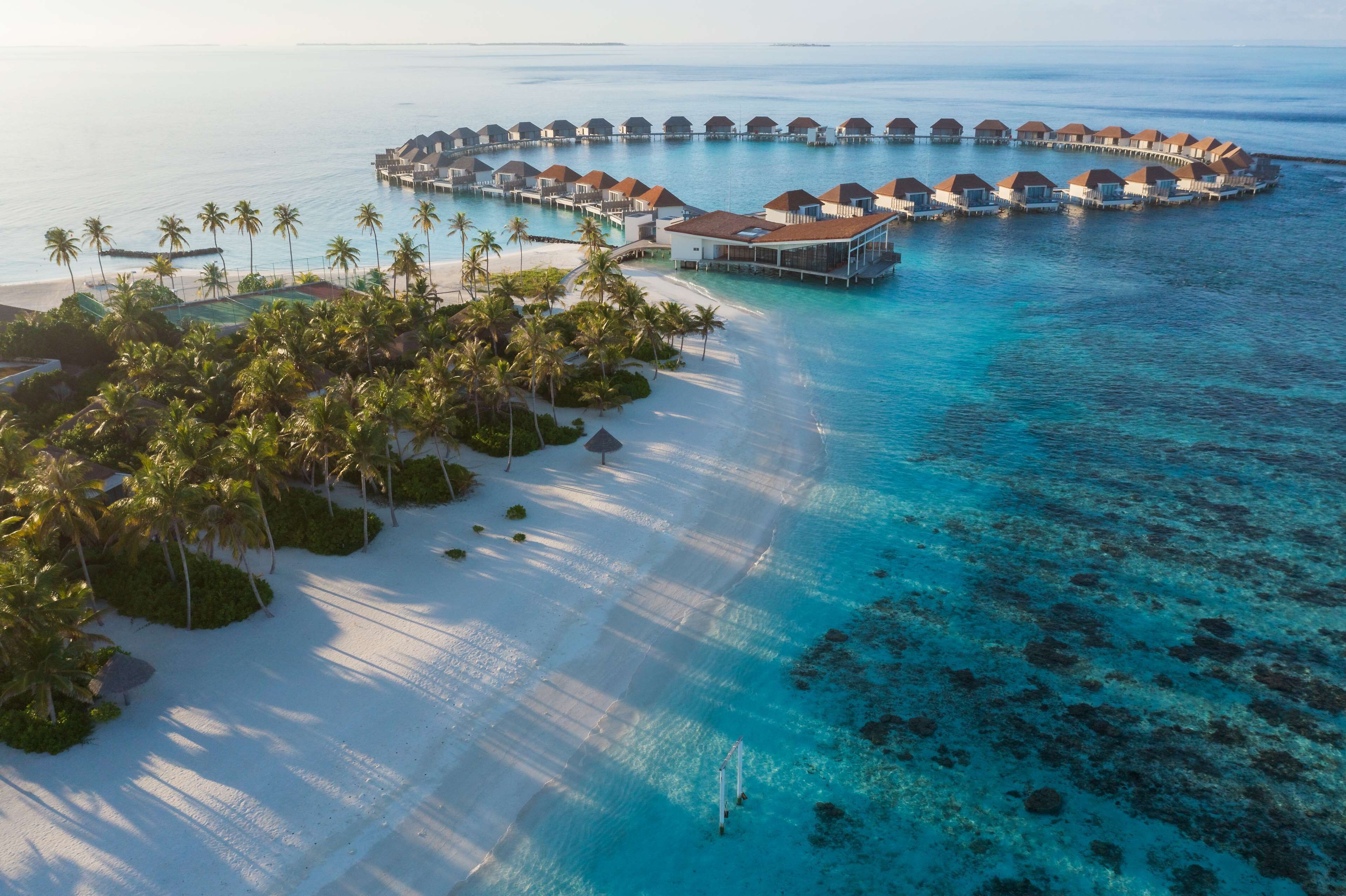 An overwater resort with multiple bungalow-style accommodations connected by walkways, a sandy beach, and palm trees.