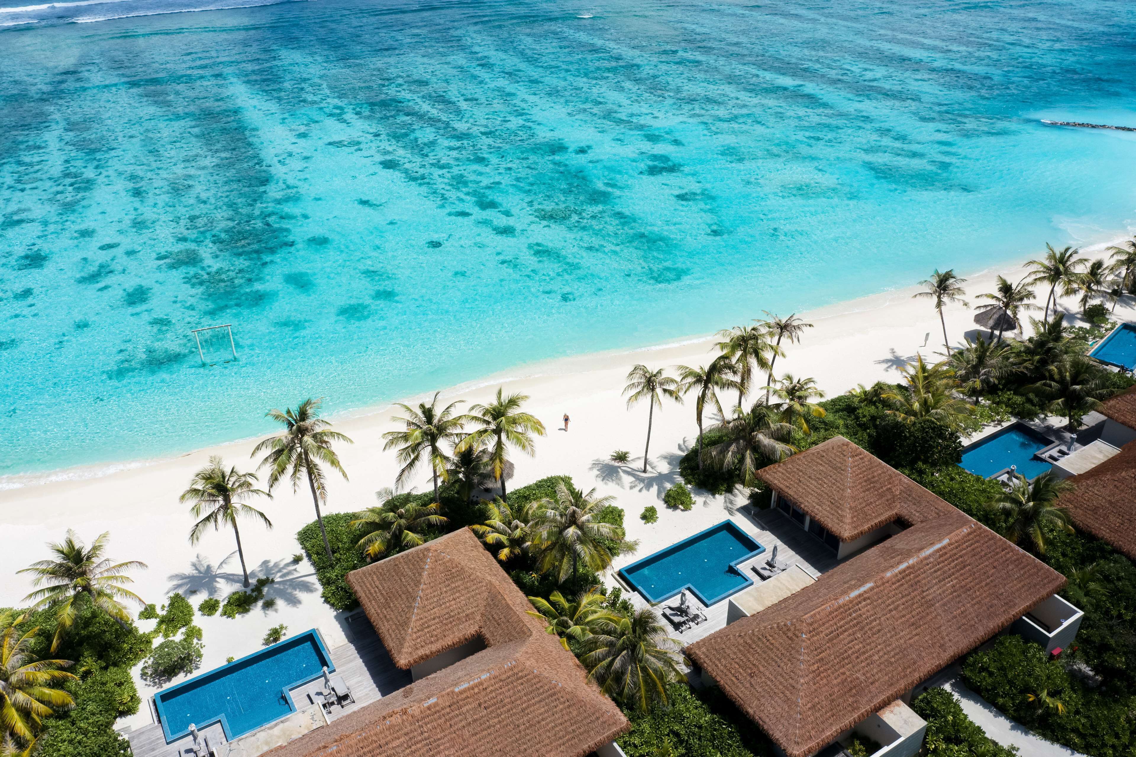 An aerial view of a resort with a clear turquoise sea, white sandy beach, and thatched-roof bungalows with swimming pools.