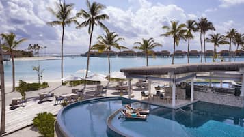 A resort pool with lounge chairs and umbrellas, surrounded by palm trees and a beach view.