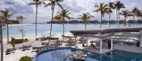 Piscina de resort con sillas de descanso y sombrillas, rodeada de palmeras y vistas a la playa.