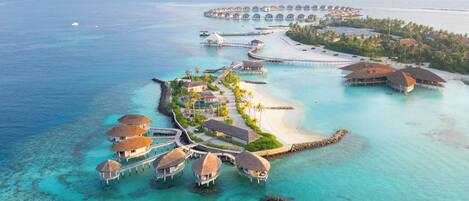 An aerial view of a resort with overwater bungalows connected by walkways, a beach, and a clear blue sea.