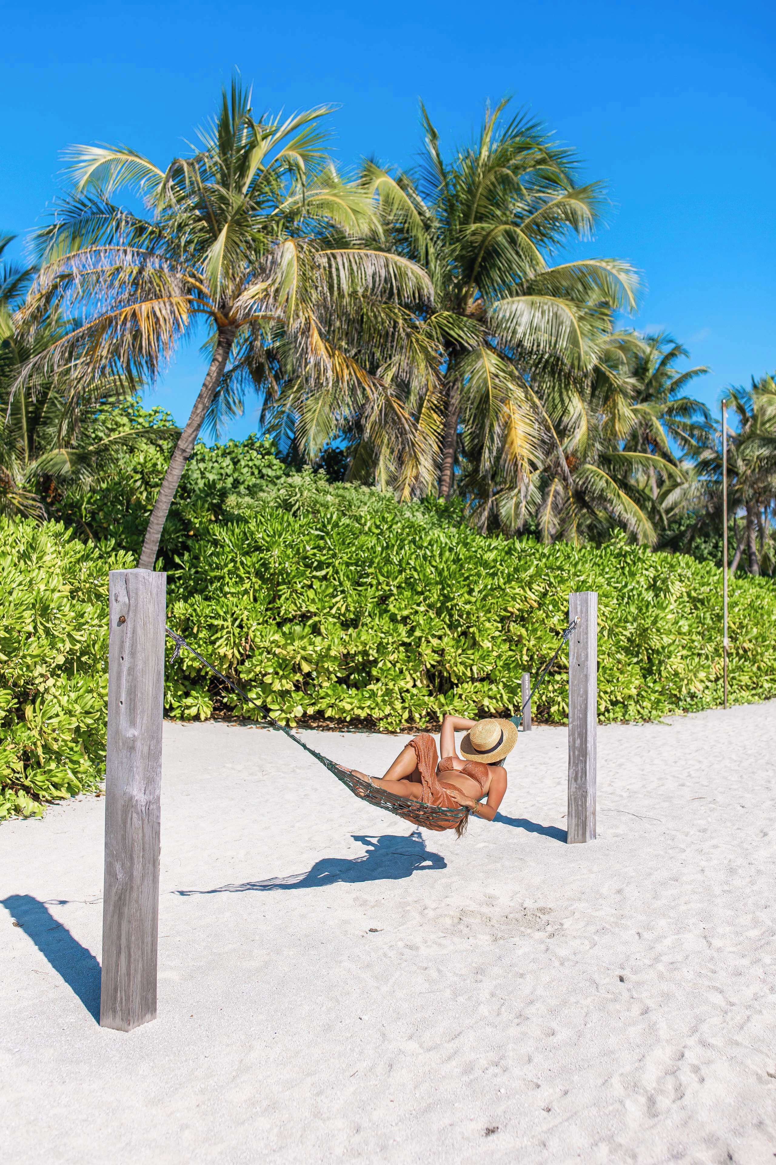 A hammock on a sandy beach with palm trees and greenery in the background.