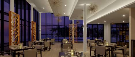 A modern dining area with tables set for two, large windows, and decorative ceiling lights.