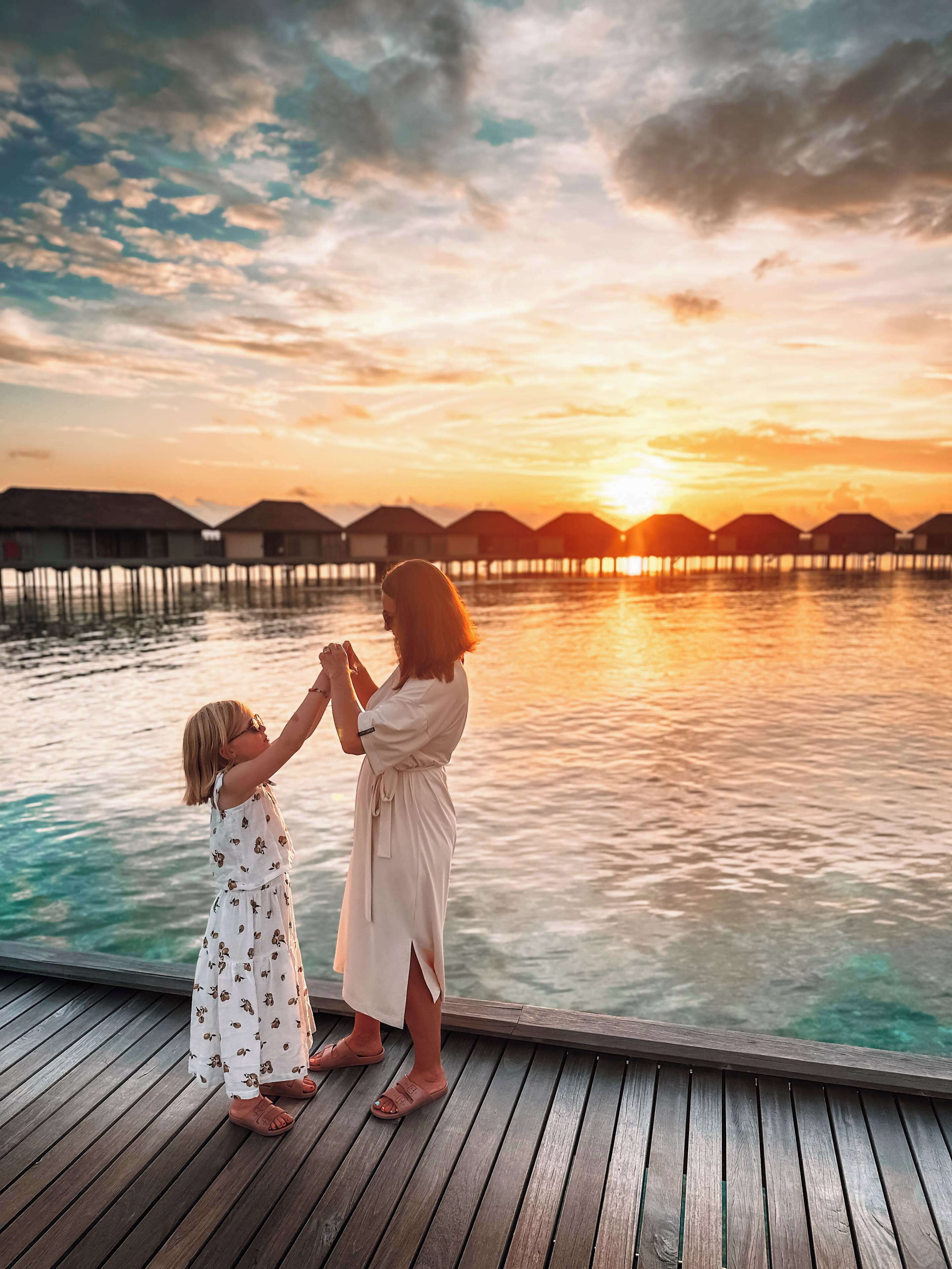 A wooden deck extending over water with overwater bungalows in the background during sunset.