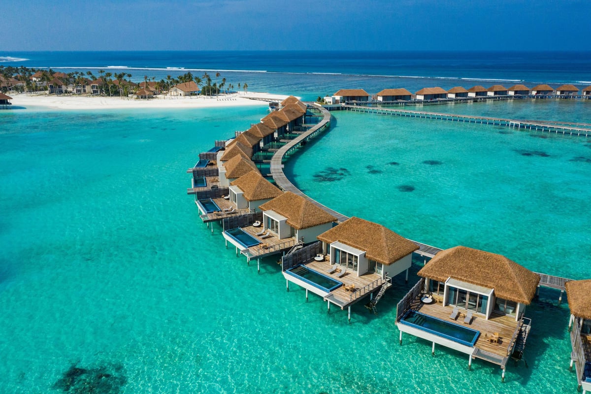 Vista aérea de bungalows sobre el agua con piscinas infinitas, unidos por un paseo marítimo, con vista a un mar turquesa y una playa de arena.