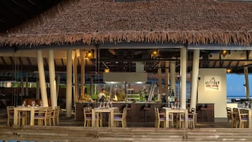 Ein Restaurant im Freien mit reetgedeckten Dächern, Holztischen und -stühlen mit Blick auf ein Gewässer.