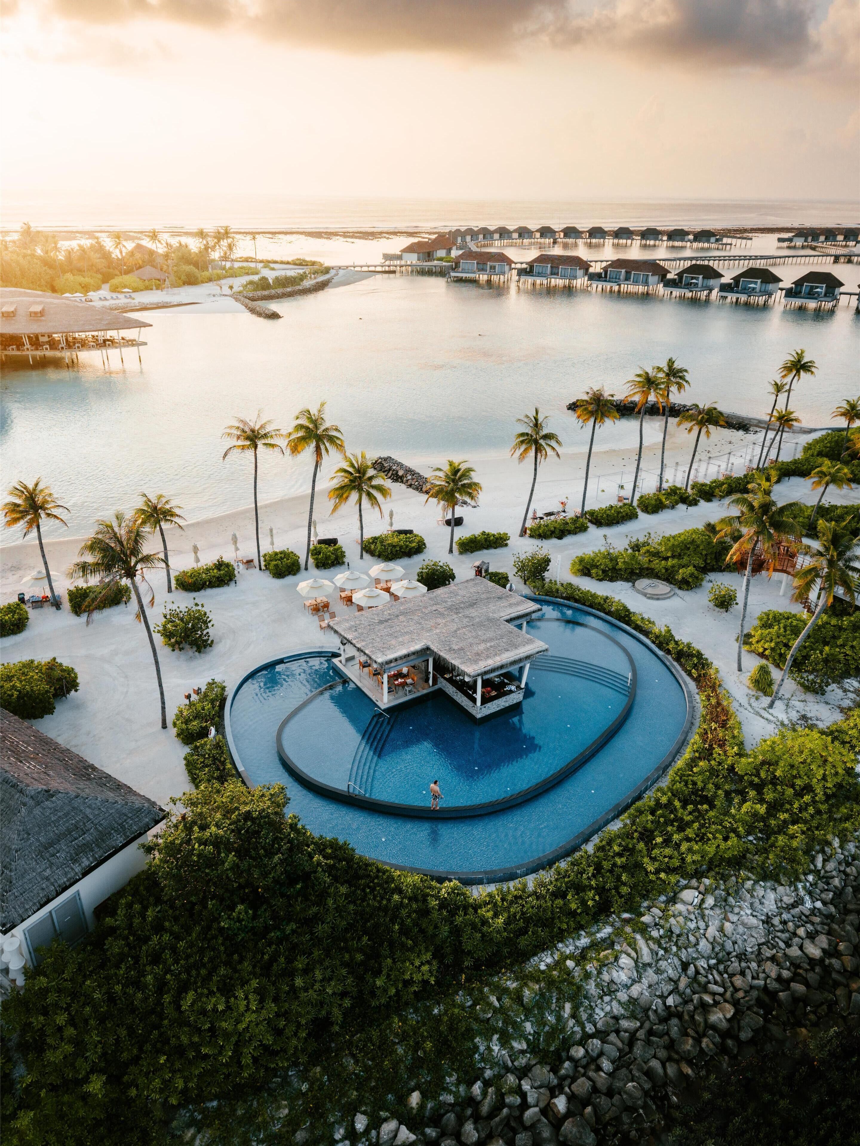 A resort with a central pool, palm trees, and overwater bungalows.