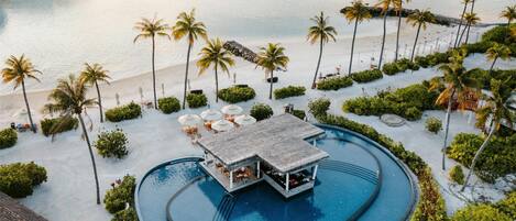 A resort with a central pool, palm trees, and overwater bungalows.