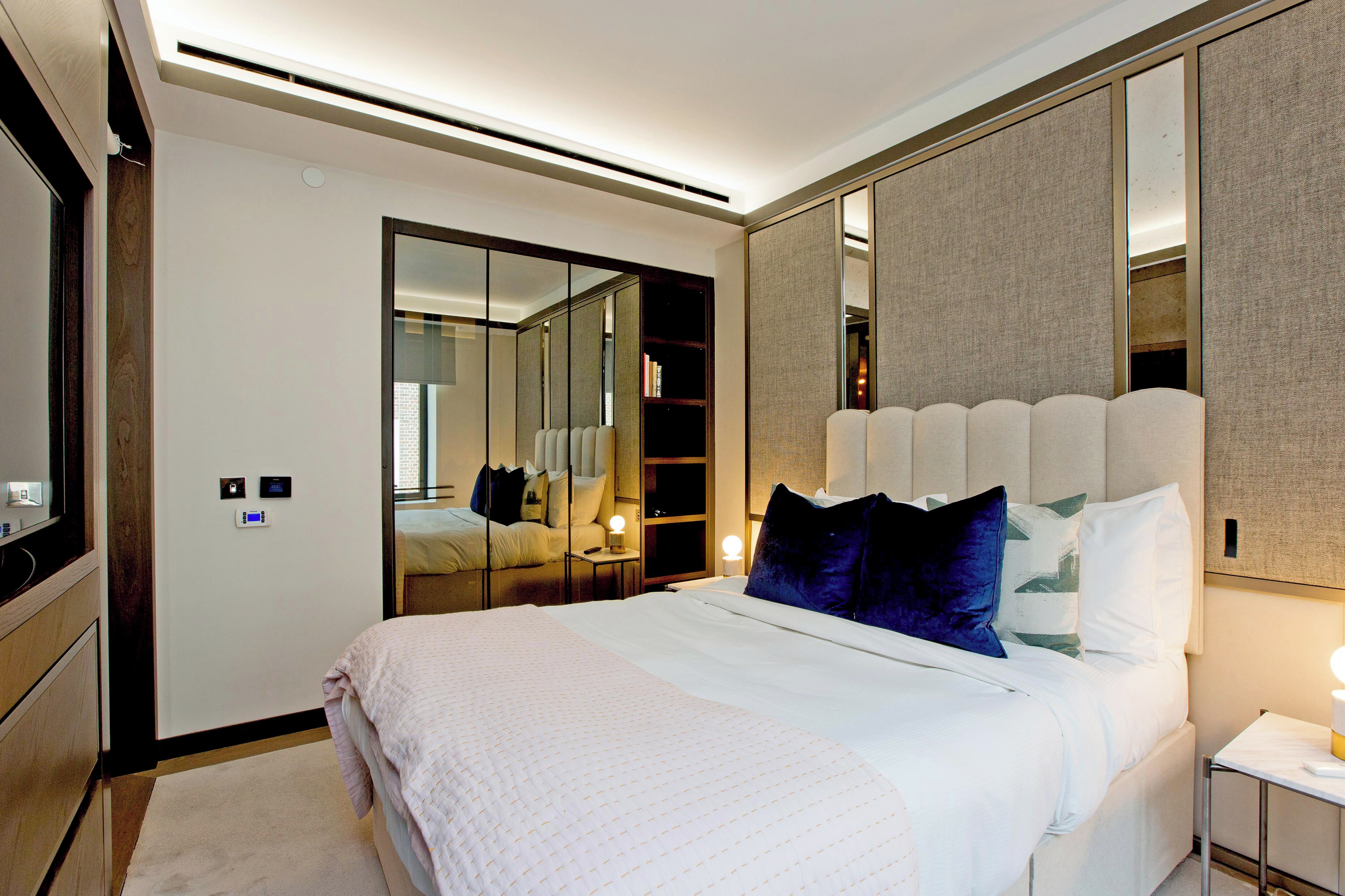 A modern hotel room with a large bed, a wardrobe, a mirror, and a bedside table with a lamp.