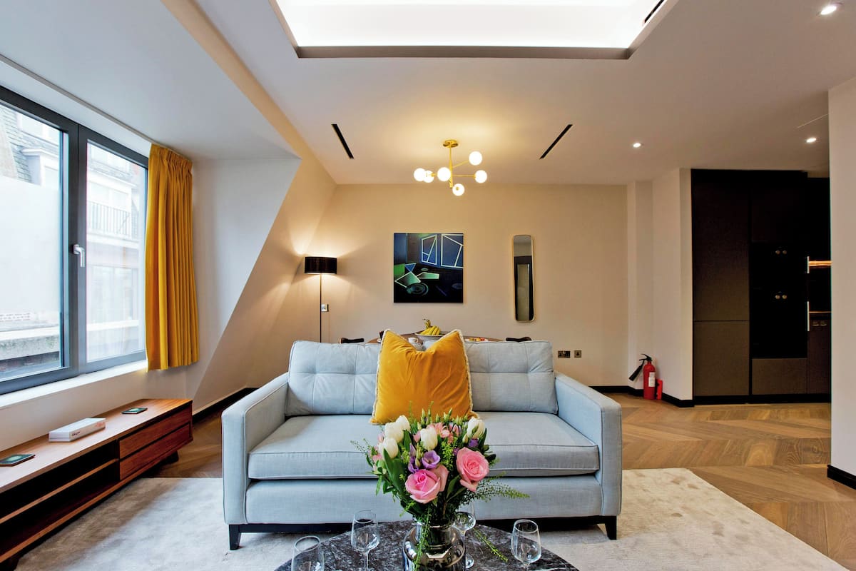 Ein modernes Wohnzimmer mit einer Couch, einem Couchtisch mit Vase und Gläsern, einem großen Fenster mit Vorhängen und einer Deckenleuchte.