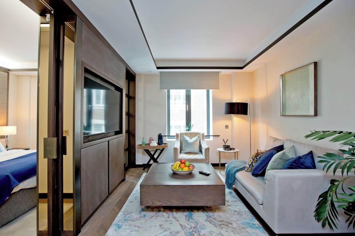 Ein modernes Hotelzimmer mit einem großen Bett, einer Couch, einem Couchtisch mit Früchten, einem Fernseher und Blick auf die Stadt.