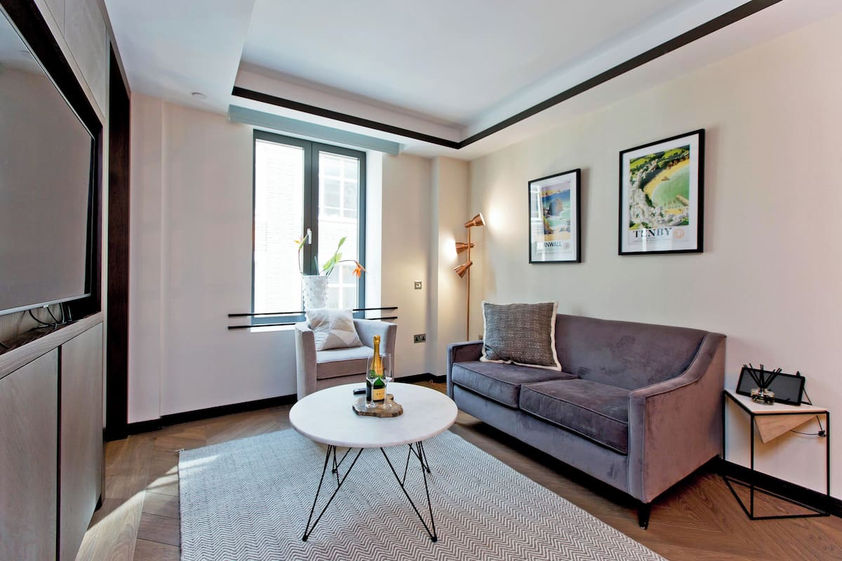 Ein modernes Wohnzimmer mit grauem Sofa, einem weißen Couchtisch mit Flasche und Glas, einem weißen Sessel und einem Fenster mit Blick.