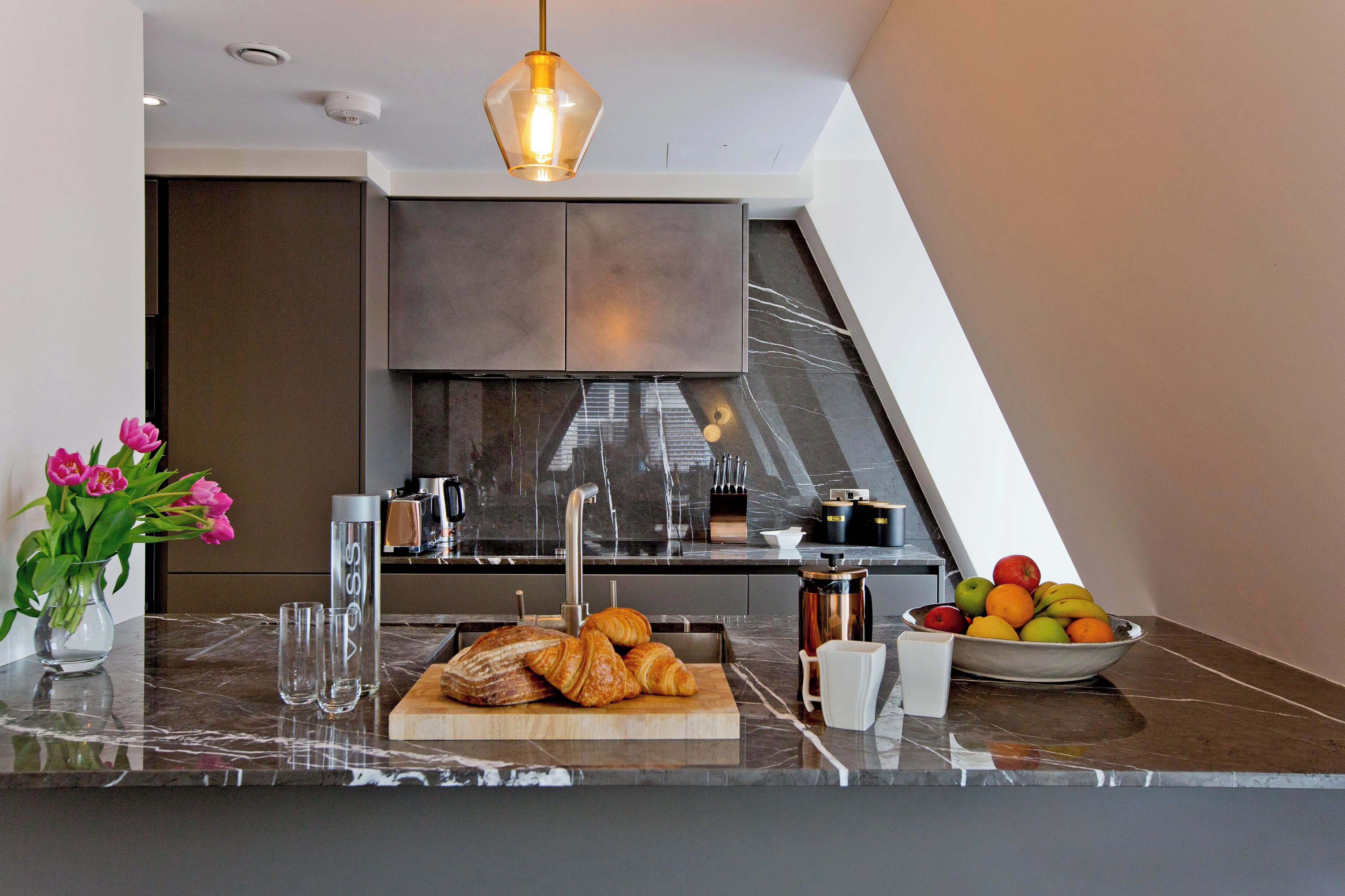 A modern kitchen with a marble countertop, a wooden cutting board with croissants, a bowl of fruit, and a vase of flowers.