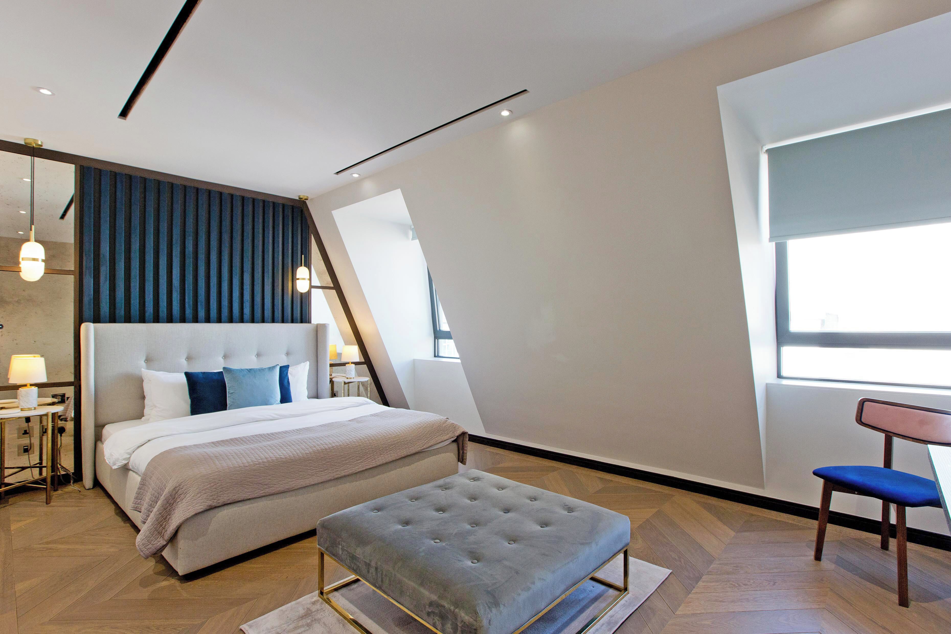 A modern bedroom with a large bed, a tufted ottoman, a chair, and a window with blinds.
