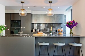 A modern kitchen with a marble countertop, two pendant lights, a built-in oven, and a bar area with stools. - 37GS Residences (London)
