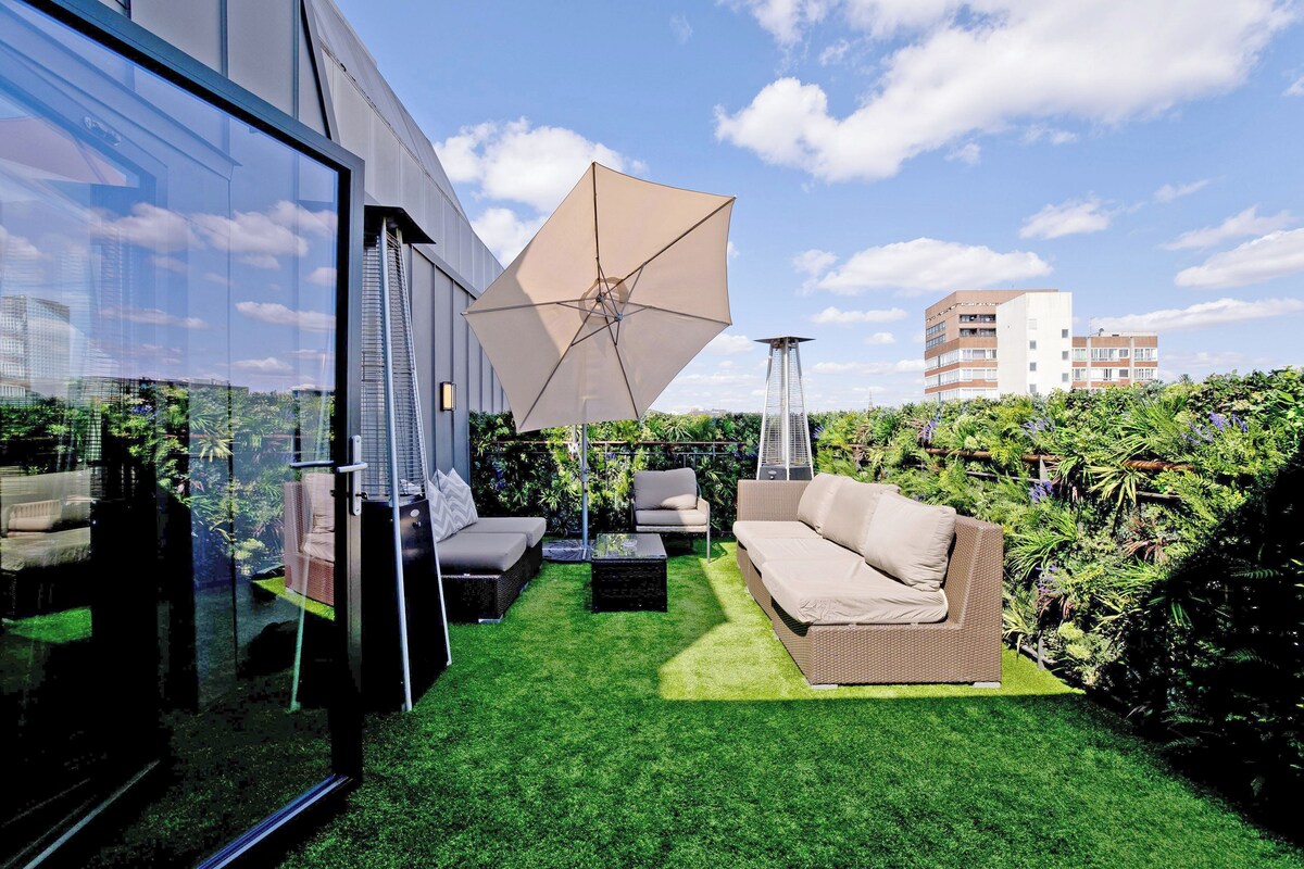 A rooftop garden with outdoor furniture, a large umbrella, and a glass wall reflecting the cityscape.