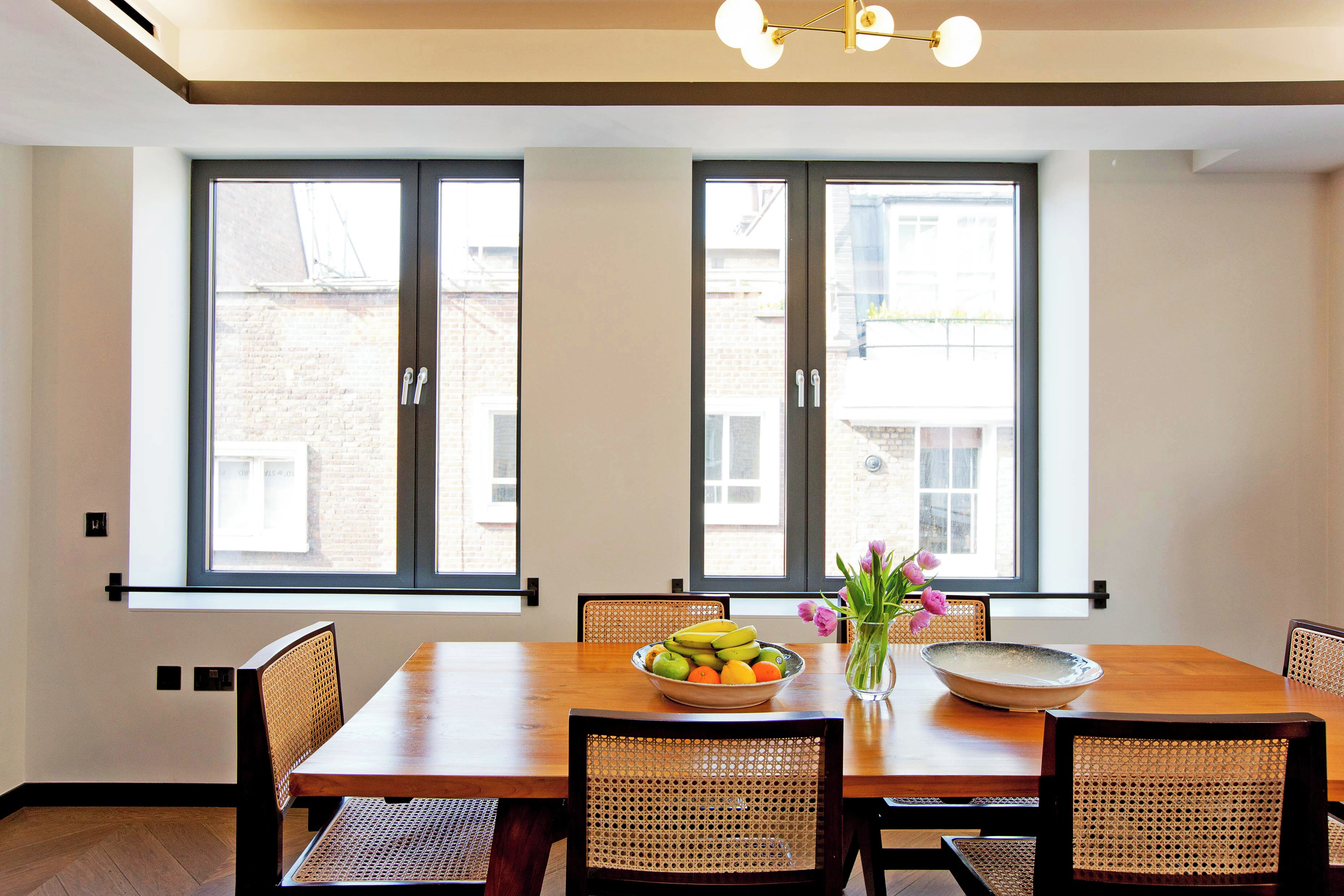 A dining area with a wooden table, wicker chairs, a bowl of fruit, and a vase with flowers.