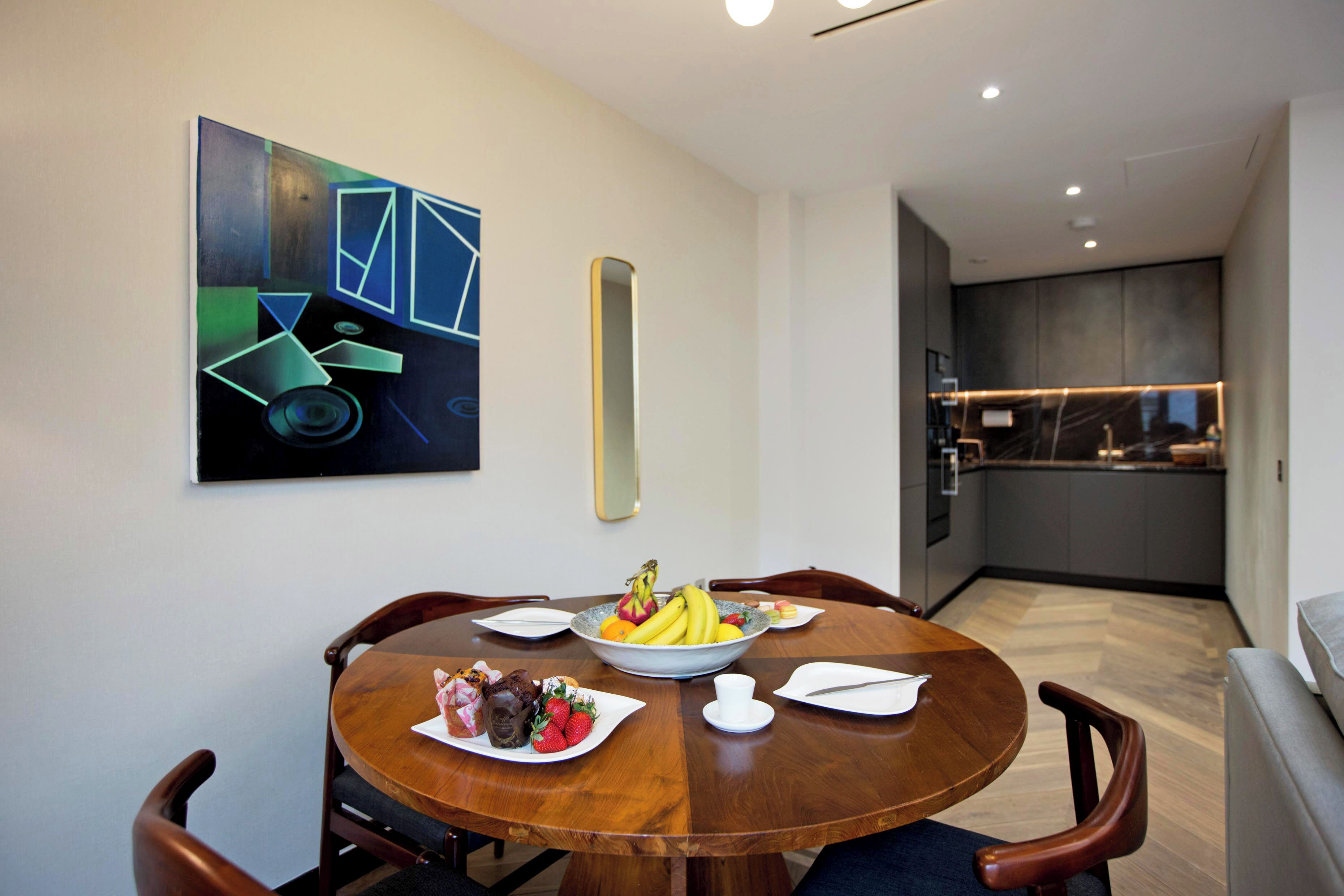 A modern dining area with a round wooden table, chairs, and a fruit bowl. A kitchen is visible in the background.